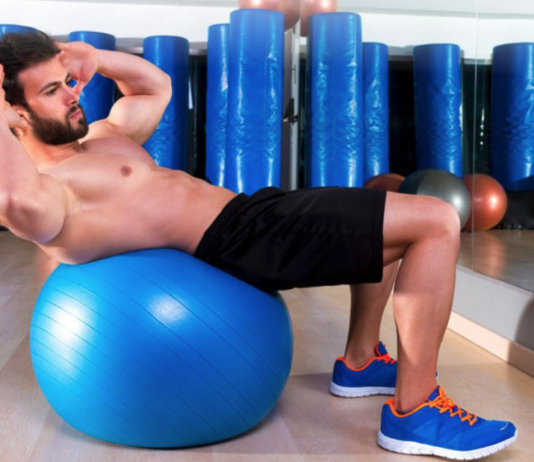 Man performing an abdominal crunch on a blue stability ball in a gym with mirrors and equipment in the background.