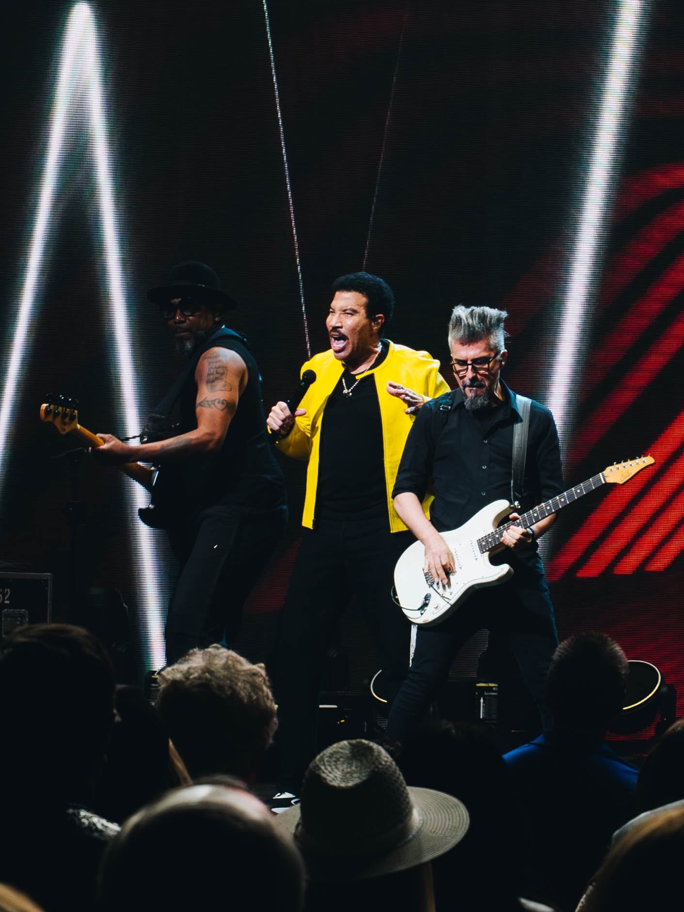 Out With Scott – By Scott Holland | Hotspots Magazine Hotspots Magazine Three musicians perform on stage: center singer in a bright yellow jacket with a microphone, flanked by guitarists on left and right against neon lights.