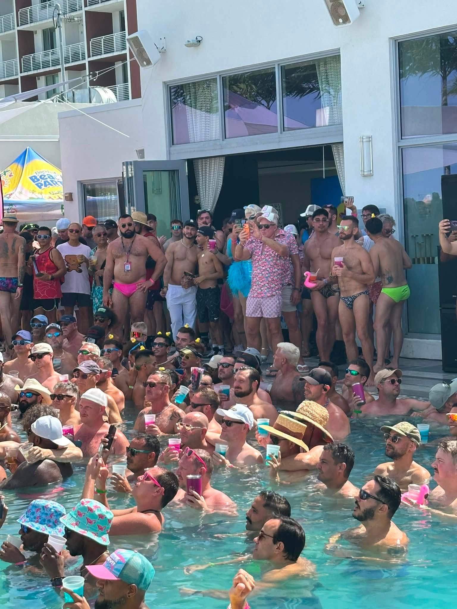 Large crowd of people in swimsuits and drinks crowding the pool area beside a modern white building, some taking photos, under sunny skies.