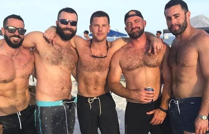 Five shirtless men stand on a sunny beach with arms around each other, wearing shorts; one holds a can and others wear sunglasses.