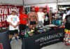 Out With Scott – With Scott Holland Five men pose at an outdoor Pride booth with rainbow bracelets and a red banner behind them promoting health care charity.