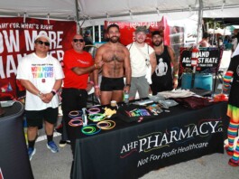Five men pose at an outdoor Pride booth with rainbow bracelets and a red banner behind them promoting health care charity.