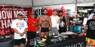 Five men pose at an outdoor Pride booth with rainbow bracelets and a red banner behind them promoting health care charity.