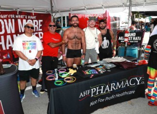Out With Scott – With Scott Holland Five men pose at an outdoor Pride booth with rainbow bracelets and a red banner behind them promoting health care charity.