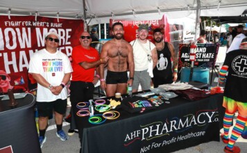 Out With Scott – With Scott Holland Five men pose at an outdoor Pride booth with rainbow bracelets and a red banner behind them promoting health care charity.
