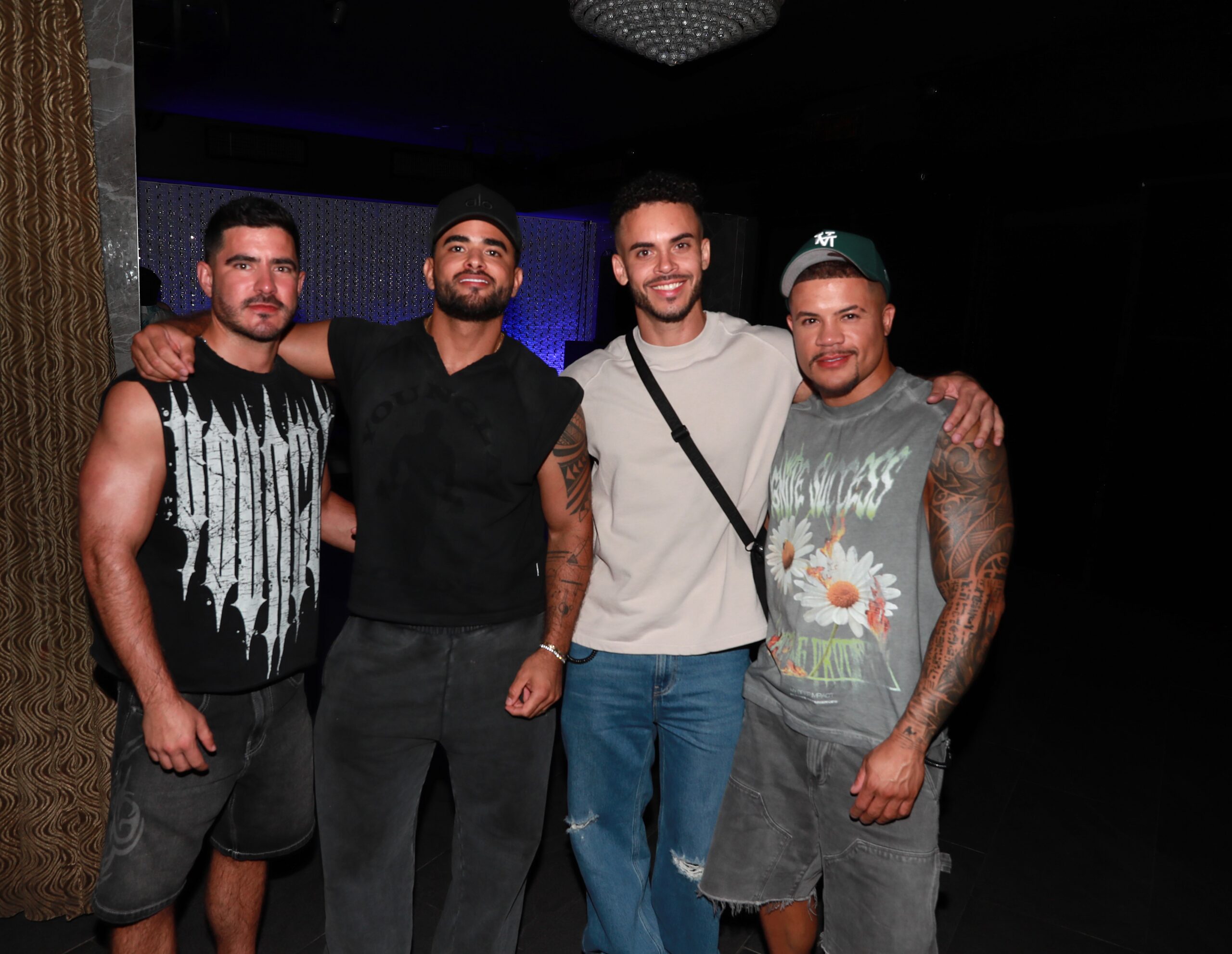 Four men pose together with arms around each other in a dim nightclub setting, smiling at the camera, casual outfits and visible tattoos on arms.