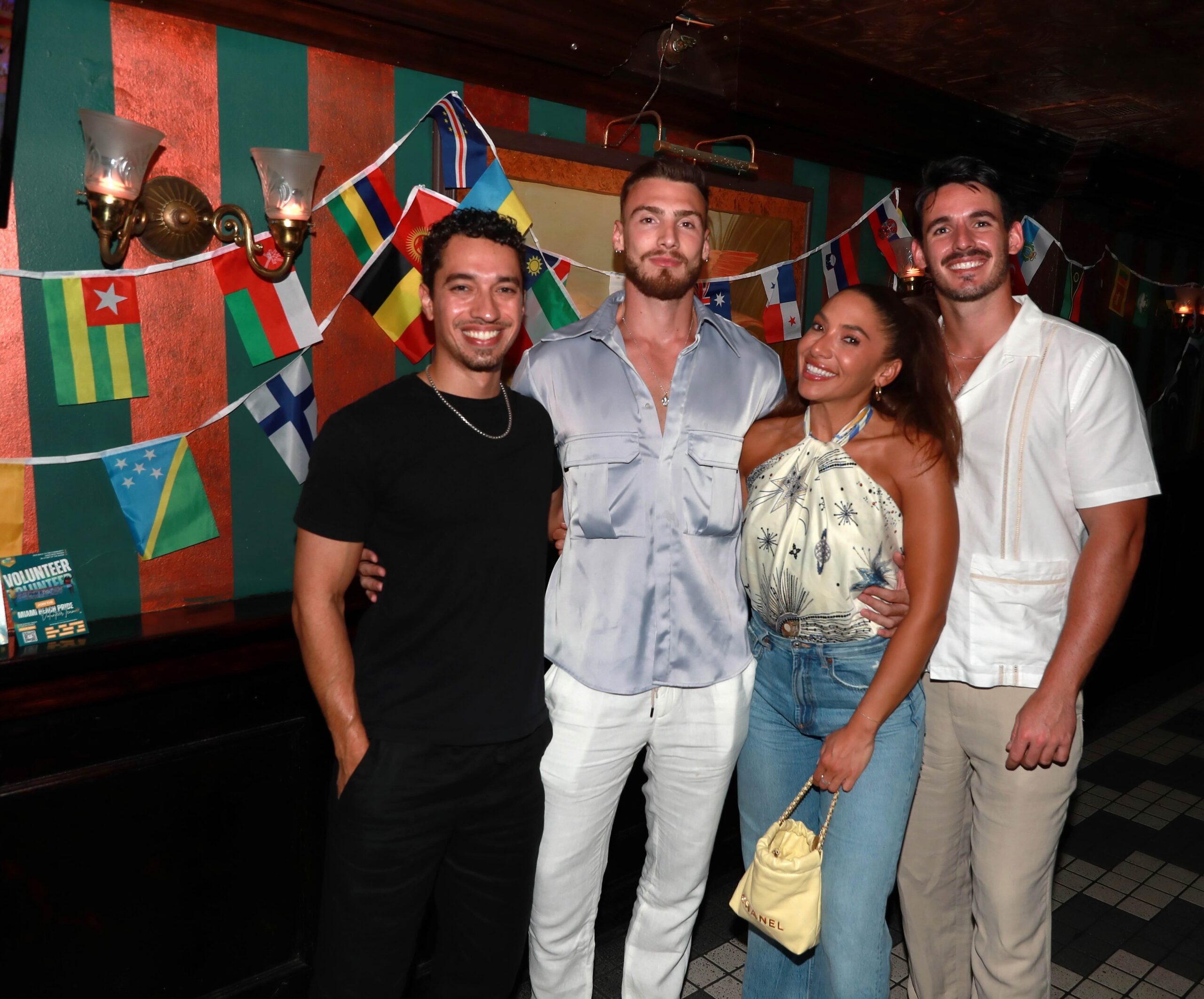 Out With Scott – By Scott Holland | Hotspots Magazine Hotspots Magazine Group of four friends posing together at a festive event with colorful flag garlands in the background.