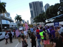 Crowd of protesters marching on a city street, many waving American flags and holding signs.