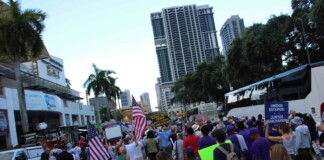 Crowd of protesters marching on a city street, many waving American flags and holding signs.