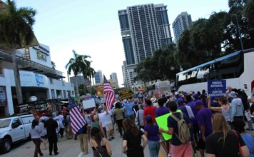Crowd of protesters marching on a city street, many waving American flags and holding signs.