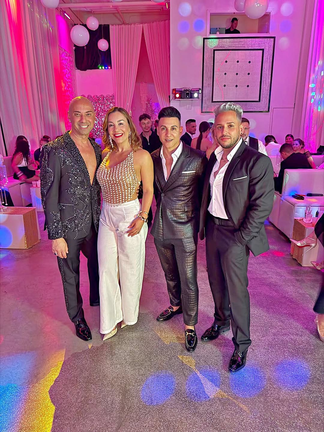 Out With Scott – By Scott Holland | Hotspots Magazine Hotspots Magazine Four people posing for a group photo at a stylish party in a pink-lit venue, wearing glam outfits including sequins and suits.