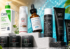Assorted skincare products on a white pedestal with a bright splashy background, featuring CeraVe cleanser, EltaMD UV Clear, Skinceuticals CE Ferulic, Paula's Choice exfoliant, and Tiëge/Brickell tubes and boxes.