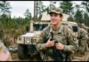 Transgender Troops Win Key Court Ruling in Military Ban Case U.S. Army soldier in camouflage holding a rifle, standing in front of a beige military vehicle in a forest clearing.