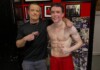 Report Says Elliot Page Will Play Sinon in Christopher Nolan’s The Odyssey Two men posing for a photo: one shirtless athlete with defined abs in red shorts, arm around a smiling trainer in a black shirt with his finger pointing at the athlete's core, against a wall covered with photos and red decor.