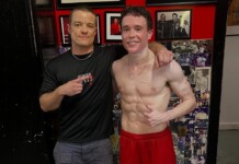 Two men posing for a photo: one shirtless athlete with defined abs in red shorts, arm around a smiling trainer in a black shirt with his finger pointing at the athlete's core, against a wall covered with photos and red decor.