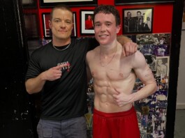 Report Says Elliot Page Will Play Sinon in Christopher Nolan’s The Odyssey Two men posing for a photo: one shirtless athlete with defined abs in red shorts, arm around a smiling trainer in a black shirt with his finger pointing at the athlete's core, against a wall covered with photos and red decor.