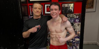Two men posing for a photo: one shirtless athlete with defined abs in red shorts, arm around a smiling trainer in a black shirt with his finger pointing at the athlete's core, against a wall covered with photos and red decor.