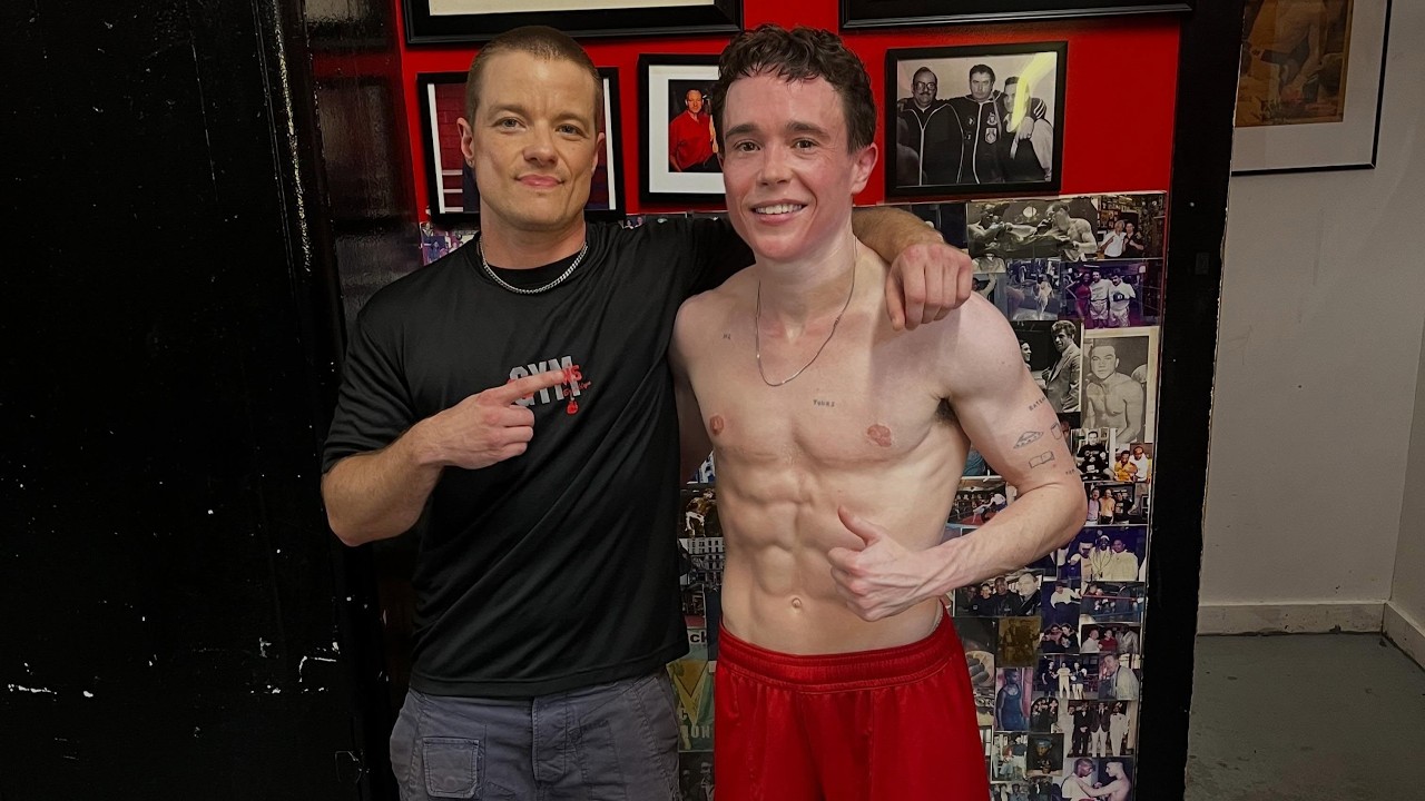 Two men posing for a photo: one shirtless athlete with defined abs in red shorts, arm around a smiling trainer in a black shirt with his finger pointing at the athlete's core, against a wall covered with photos and red decor.