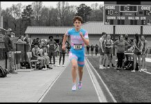 Runner in a blue-pink uniform sprinting on a track with bib 102, as spectators watch from the sidelines.
