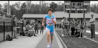 Runner in a blue-pink uniform sprinting on a track with bib 102, as spectators watch from the sidelines.
