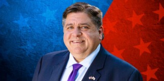 Portrait of a smiling man in a dark suit and purple tie, wearing a US flag lapel pin, against a blue and red starry background.