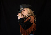 After Everything, Melissa Etheridge Is Still Rising Woman wearing a black cowboy hat with a decorative band and a brown fringe jacket, hands pressed together in a prayer pose against a black backdrop.