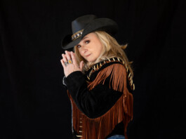 After Everything, Melissa Etheridge Is Still Rising Woman wearing a black cowboy hat with a decorative band and a brown fringe jacket, hands pressed together in a prayer pose against a black backdrop.