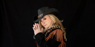 Woman wearing a black cowboy hat with a decorative band and a brown fringe jacket, hands pressed together in a prayer pose against a black backdrop.