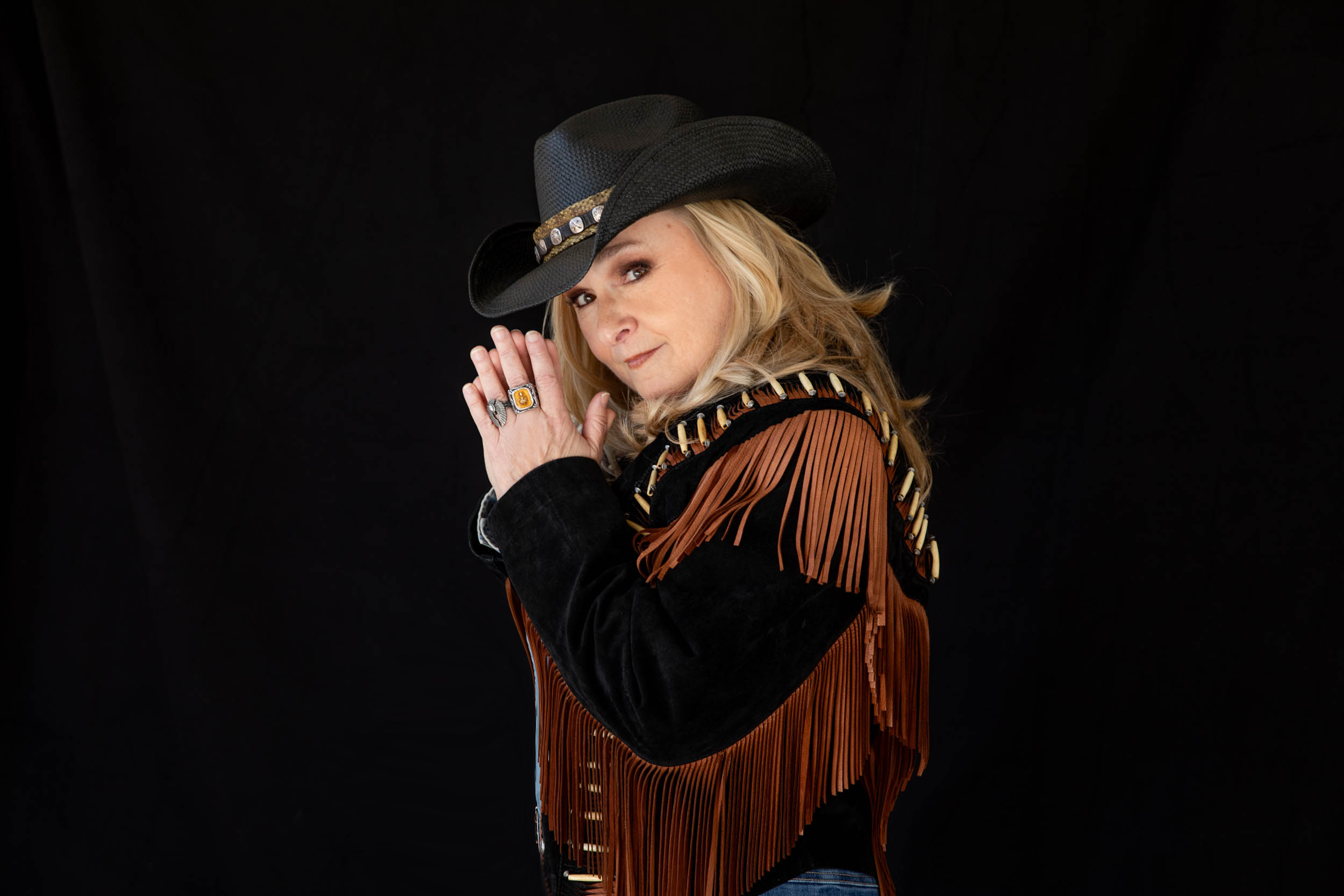 Photographed in Joshua Tree California October 22 2025 Woman wearing a black cowboy hat with a decorative band and a brown fringe jacket, hands pressed together in a prayer pose against a black backdrop.