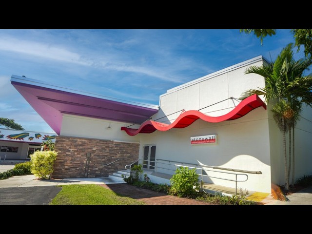 Exterior of a white commercial building with a red wavy canopy, brick accents, and ramp access under a blue sky.
