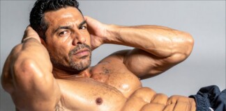 Shirtless muscular man, hands behind head, showing defined abs in a studio portrait.