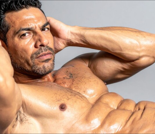 Shirtless muscular man, hands behind head, showing defined abs in a studio portrait.