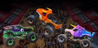 Three colorful monster trucks mid-air over a dirt arena as a crowd watches a high-energy show.