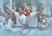 Two men in white T-shirts ride a boat and wave to a cheering crowd at a sunny parade.