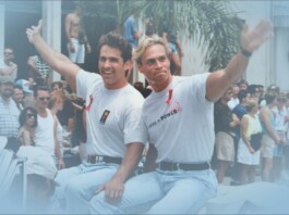 Deep Inside Hollywood: A Bloody Prom, Queer Bodybuilders, and the Return of the ’90s Supers Two men in white T-shirts ride a boat and wave to a cheering crowd at a sunny parade.