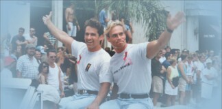 Two men in white T-shirts ride a boat and wave to a cheering crowd at a sunny parade.