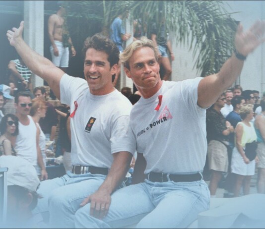 Two men in white T-shirts ride a boat and wave to a cheering crowd at a sunny parade.
