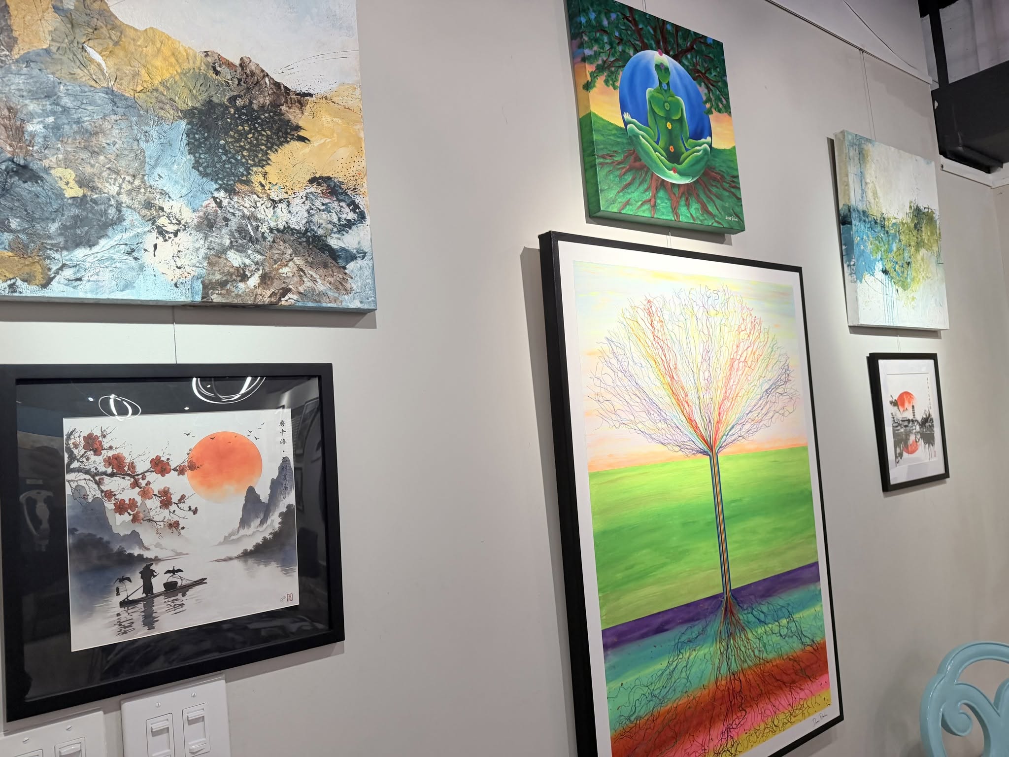 Gallery wall with several colorful paintings: left abstract blue-yellow piece, center-top green meditating figure under a tree, and a rainbow-tree work below.