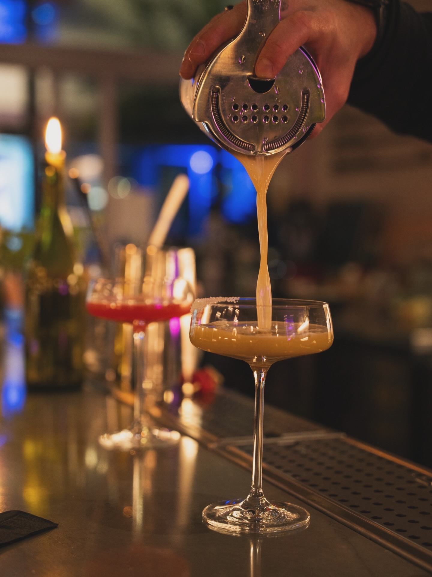 Cocktail being poured from a metal shaker into a coupe glass at a bar, with bottles in the blurred background