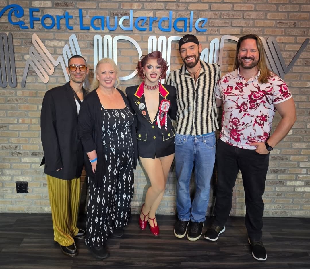 Group of five people posing together in front of a brick wall with a Fort Lauderdale sign visible in the background; one person in the center wears a bold jacket, red accessories, and heels, resembling a drag performance costume.