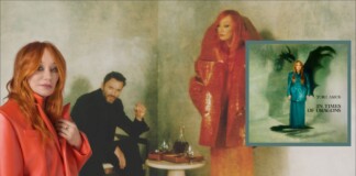 Red-haired woman in an orange coat stands at left; a man in black sits behind beside a table with glasses, against a moody green backdrop; an inset album cover appears on the right.
