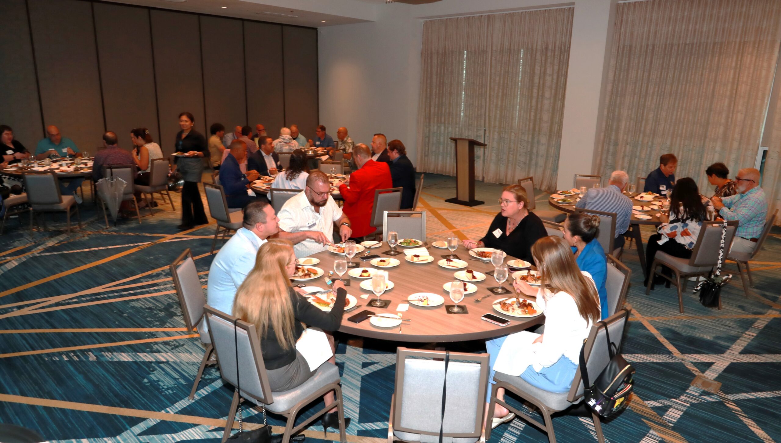 Group of people seated at round tables eating during a formal dinner in a conference room in a hotel banquet setting.