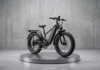 Black electric bike on a round metallic pedestal in a minimalist concrete studio setting.