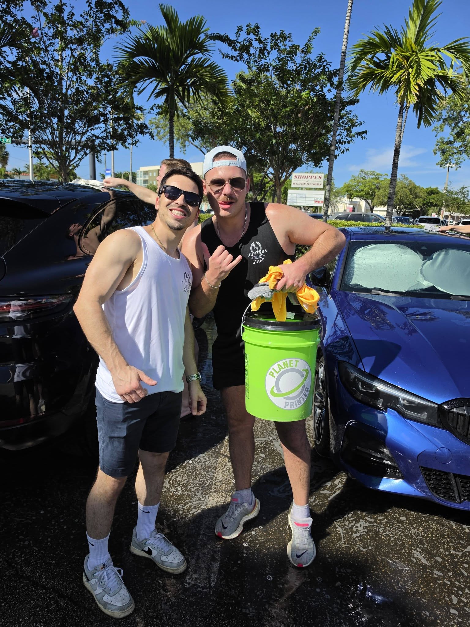 Two young men posing for a photo by parked cars, one holding a bright green Planet Printer bucket with towels. The scene appears to be at a car wash or outdoor event.
