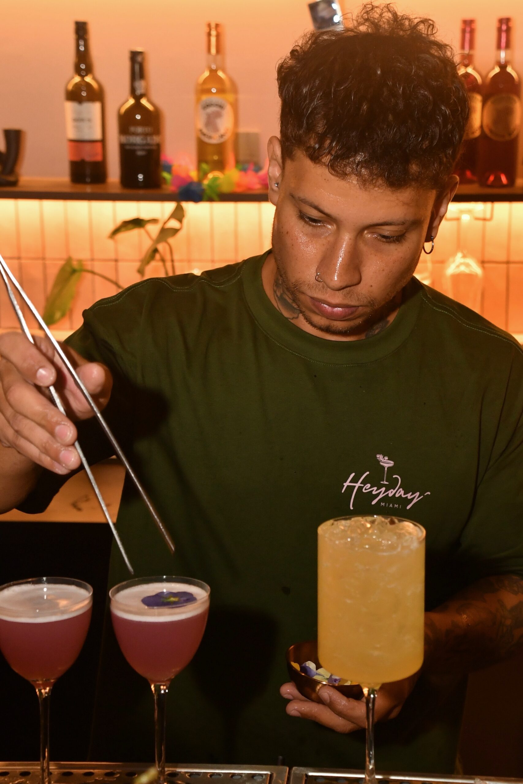 Bartender in a green shirt preparing cocktails at a bar, with three pink drinks and a tall yellow cocktail, liquor bottles on a backlit shelf behind.