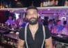 Bearded bartender wearing a black shirt and suspenders behind a busy bar.