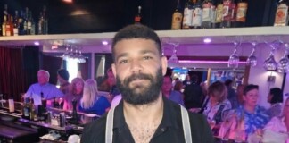 Bearded bartender wearing a black shirt and suspenders behind a busy bar.