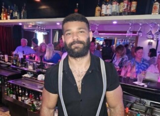 Bearded bartender wearing a black shirt and suspenders behind a busy bar.