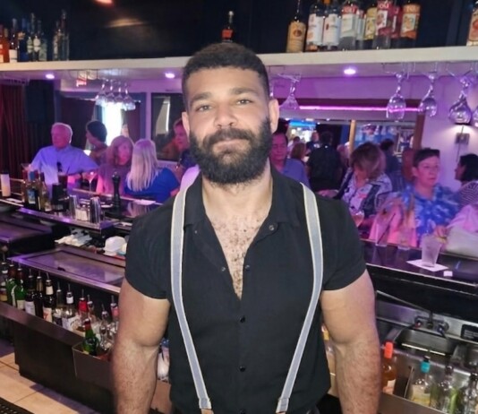 Bearded bartender wearing a black shirt and suspenders behind a busy bar.
