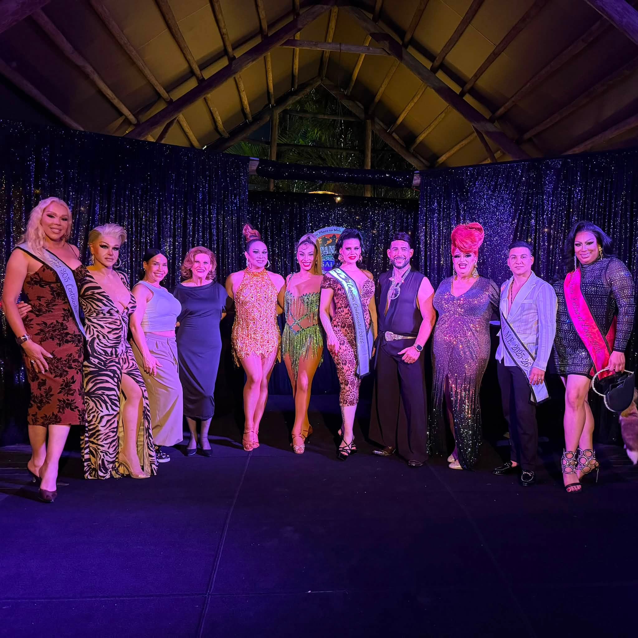 Group of drag pageant contestants posing on stage in glamorous dresses with sashes, under twinkling purple lights.